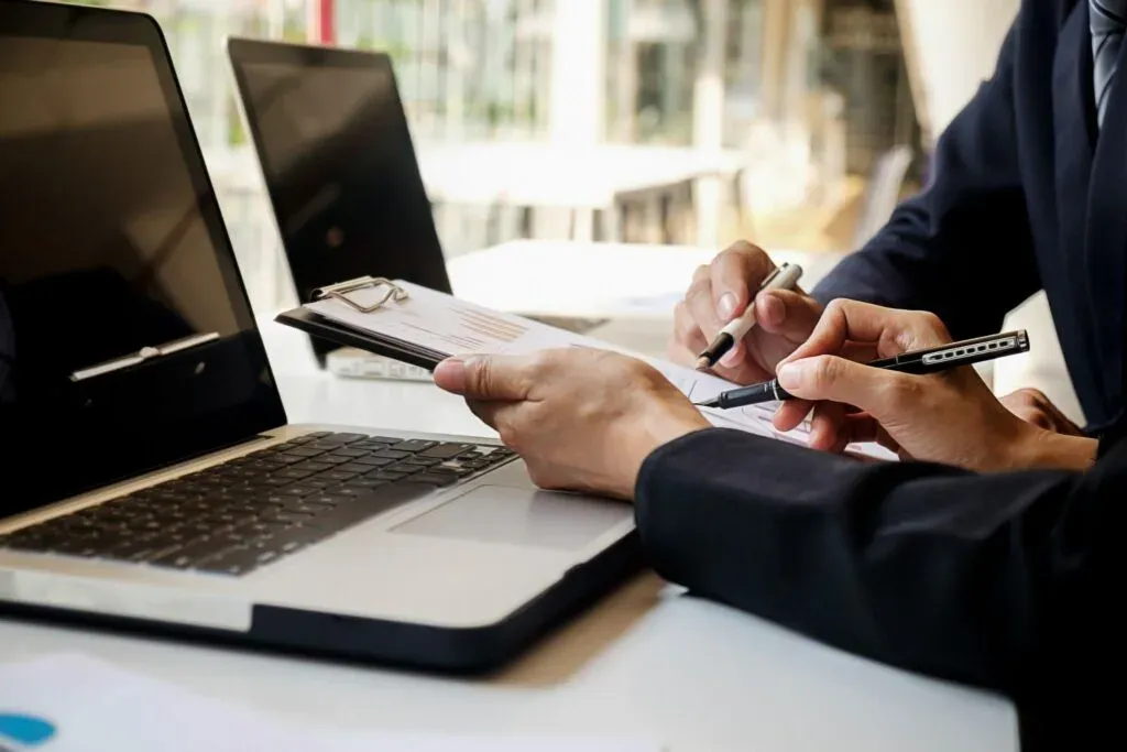 Close-up of two people at a laptop, one signing a document on a clipboard.