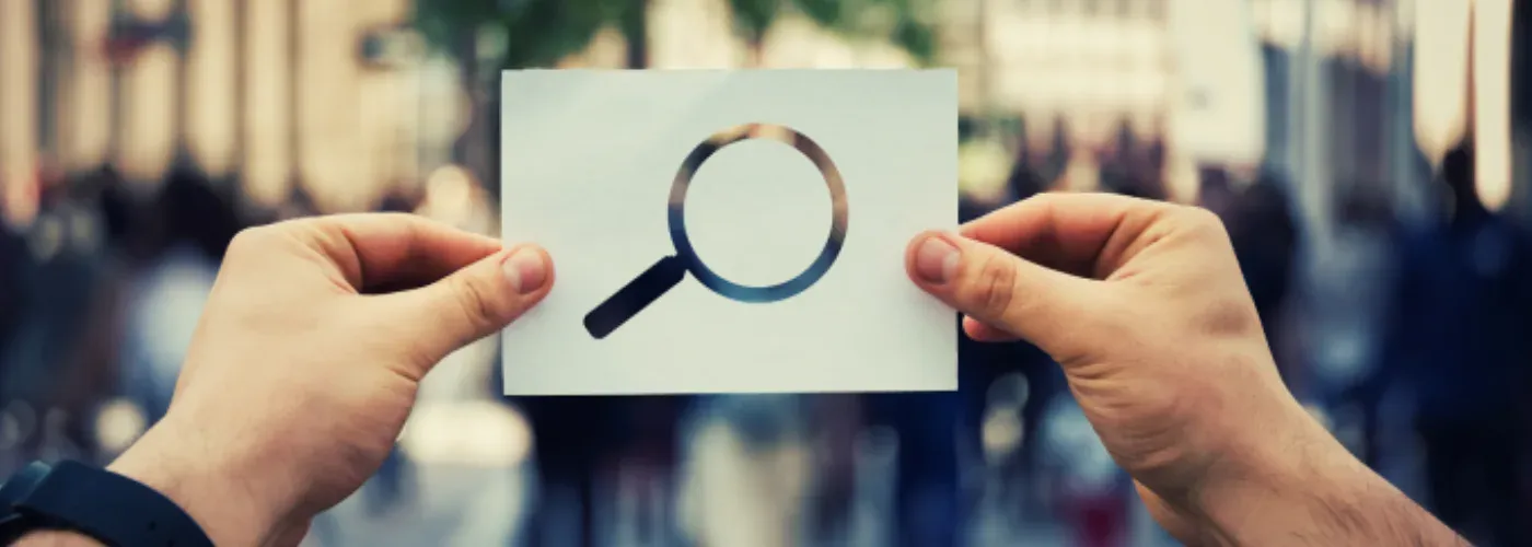 Two hands holding up a card printed with a magnifying-glass search icon against a blurred street.