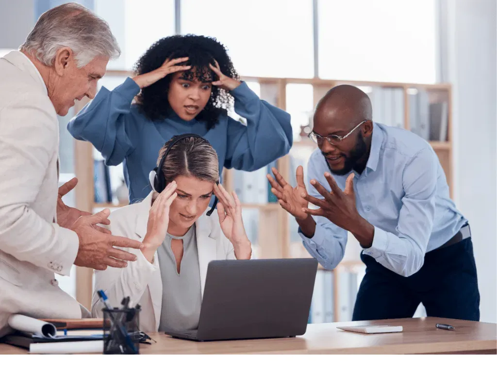 Four colleagues clustered around a laptop, looking stressed and frustrated.