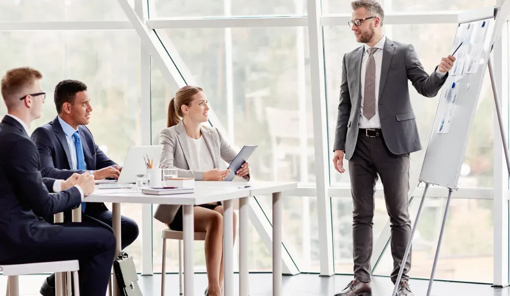 A presenter at a flip chart leading three seated colleagues through a training session in a bright meeting room.